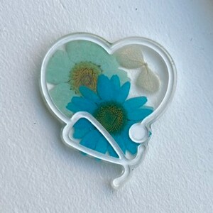 Pode incluir: Um ornamento de resina branco em forma de cora&ccedil;&atilde;o com um design de estetosc&oacute;pio e flores prensadas dentro. As flores s&atilde;o azuis, brancas e verdes.