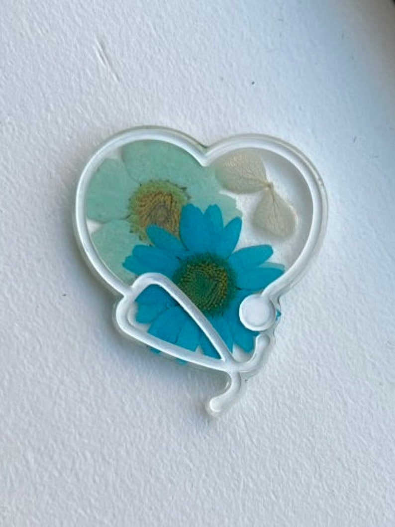 Pode incluir: Um ornamento de resina branco em forma de cora&ccedil;&atilde;o com um design de estetosc&oacute;pio e flores prensadas dentro. As flores s&atilde;o azuis, brancas e verdes.