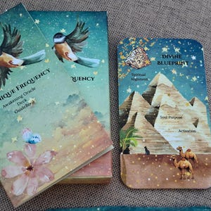 May include: A rectangular oracle deck box and a single card featuring a starry night sky. The box displays a bird, a flower, and the text Unique Frequency Awakening Oracle Deck GuideBook. The card shows pyramids, camels, and the text Divine Blueprint.
