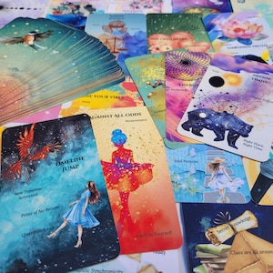May include: A deck of oracle cards featuring colorful illustrations and text. Visible cards include 'Timeline Jump' with a person in a blue dress, 'Against All Odds' with a figure in a red dress, and 'Divine Timing' with a bear and celestial imagery.