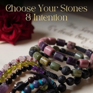 May include: An assortment of gemstone bracelets in various colors, including pink, green, purple, and black, are displayed. A red rose and the text "Choose Your Stones & Intention" are in the background. The bracelets are on a light-colored surface.