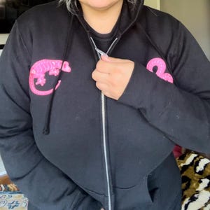 Puede incluir: Sudadera negra con cremallera y diseño de gecko rosa en el pecho y la manga. La sudadera tiene una capucha con cordón y una cremallera plateada. El diseño del gecko es de color rosa brillante.