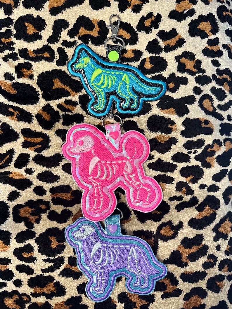 May include: A vertical chain of three embroidered dog-shaped keychains. The top keychain is blue with a neon green skeleton, the middle is pink with a white skeleton and heart, and the bottom is purple with a white skeleton and heart.