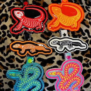 May include: A collection of embroidered keychains featuring reptile and amphibian designs. The keychains include a red lizard skeleton, an orange lizard, a black and white lizard skeleton, a blue and green snake, and a pink and yellow snake.