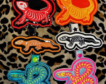 Op de afbeelding: Een verzameling geborduurde sleutelhangers met reptielen- en amfibieontwerpen. De sleutelhangers omvatten een rood hagedisskelet, een oranje hagedis, een zwart-wit hagedisskelet, een blauw-groene slang en een roze-gele slang.