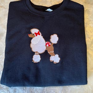May include: Black sweatshirt with an embroidered white poodle wearing a red bow and a candy cane. The poodle is brown on the bottom half of its body.