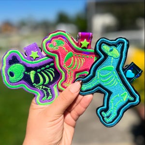May include: Three embroidered dog-shaped keychains featuring skeleton designs. The left keychain is purple with a green skeleton, the center is pink with a green skeleton, and the right is blue with a green skeleton. Each has a small charm attached.