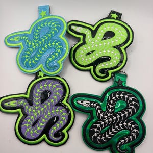 May include: Four embroidered snake-shaped keychains with star-shaped accents at the top. The snakes feature skeletal patterns in lime green or white thread on blue, lime green, purple, and black backgrounds with glossy, iridescent finishes.