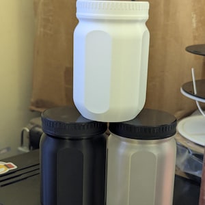 Puede incluir: Tres tarros de vidrio con tapas, uno blanco, uno negro y uno esmerilado. Los tarros están apilados uno encima del otro.