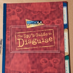 Könnte beinhalten: Ein rotes und weinrotes Buch mit dem Titel "The Spy's Guide to Disguise" mit dem Logo "SPY University". Das Buch hat Registerkarten mit der Aufschrift "Spyquest", "SPYmissions" und "SPYfeature". Das Cover zeigt Illustrationen zum Thema Spionage.