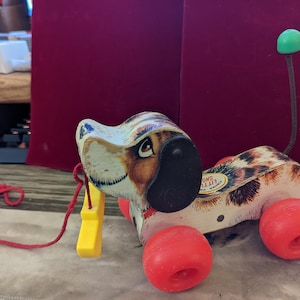 以下が含まれることがあります： 茶色、黒、白の斑点模様のついた、ヴィンテージの木製引きずりおもちゃの犬。犬は赤い車輪、黄色の引き紐、緑色の尻尾が付いています。おもちゃには「Little Snoozer」とラベルが貼られています。