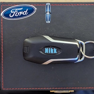 Puede incluir: Llave de coche negra y plateada con el nombre "Nikk" en azul claro. La llave está sobre una superficie de cuero negro con costuras rojas. El logotipo de Ford está en la esquina superior izquierda.