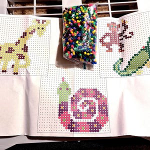 Puede incluir: Un conjunto de cuatro patrones de arte con cuentas que representan una jirafa, un caracol, una oruga y una flor. Los patrones están impresos en papel blanco con una cuadrícula de cuadrados. Las cuentas no están incluidas.