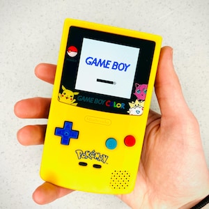 Puede incluir: Una consola de videojuegos portátil Game Boy Color amarilla con un logotipo de Pokemon y una pantalla en blanco y negro que muestra "GAME BOY".