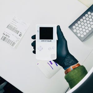 Op de afbeelding: Een witte Game Boy Color handheld spelcomputer met een zwart scherm, een richtingspad, twee ronde knoppen en een luidsprekerrooster. Het apparaat toont de tekst GAME BOY COLOR onder het scherm en heeft een klein aan-uit indicatielampje.