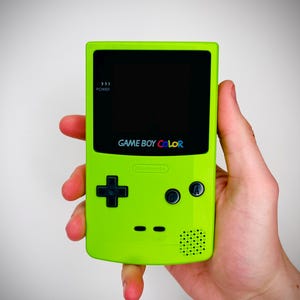 以下が含まれることがあります： ライムグリーンのゲームボーイカラー携帯ゲーム機。黒い画面ベゼル、十字キー、Aボタン、Bボタンを備え、画面の下にはGAME BOY COLORの文字がプリントされています。本体には電源インジケーターライトとスピーカーが搭載されています。