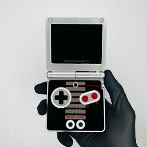 May include: Silver Game Boy Advance SP handheld game console with a black screen and a black and gray controller design. The controller has red A and B buttons, a gray cross-shaped directional pad, and the text "Nintendo".