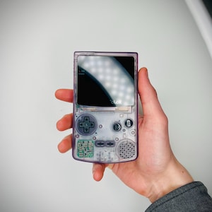 Puede incluir: Una consola de videojuegos portátil con una carcasa de plástico morado translúcido. El dispositivo cuenta con una pantalla negra, un pad direccional, botones A y B, botones de inicio y selección, y una rejilla de altavoz. La palabra COLOR está impresa.