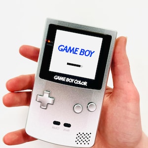 Op de afbeelding: Een zilveren Game Boy Color draagbare videogameconsole met een blauw "GAME BOY"-logo op het scherm. De console heeft een zwart-witte directionele pad, twee ronde knoppen en een luidspreker.