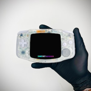 May include: A handheld Game Boy Advance video game console with a transparent clear plastic shell, white directional pad and buttons, and a black screen bezel featuring the Game Boy Advance logo in colourful text at the bottom centre.