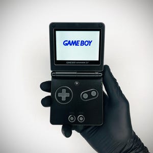 Op de afbeelding: Een zwarte Game Boy Advance SP handheld spelcomputer met een clamshell-ontwerp. Het scherm toont het blauwe Game Boy-logo. Het apparaat is voorzien van een richtingspad, A- en B-knoppen en twee kleine ronde knoppen onder het scherm.