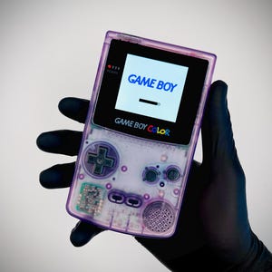 May include: A translucent purple handheld gaming console with a black screen bezel. The screen displays the text GAME BOY. Below the screen, the text GAME BOY COLOR is printed. The device features a directional pad, A and B buttons, and a speaker.
