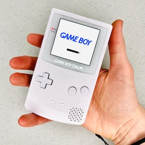 Puede incluir: Una consola de videojuegos portátil Game Boy Color blanca con un logotipo "GAME BOY" azul en la pantalla. La consola está en la mano de una persona.