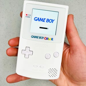 Könnte beinhalten: Eine weiße Game Boy Color Handheld-Konsole. Der Bildschirm zeigt "GAME BOY" in blauer Schrift. Die Konsole hat ein Steuerkreuz, A- und B-Tasten und einen Lautsprecher. Der Text "GAME BOY COLOR" ist unter dem Bildschirm aufgedruckt.