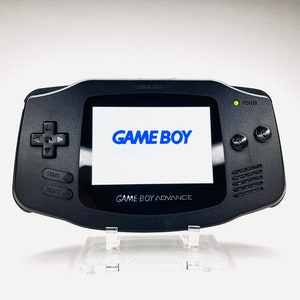 Puede incluir: Una consola de videojuegos portátil Game Boy Advance negra con un logotipo "GAME BOY" azul en la pantalla. La consola está en un soporte de plástico transparente.