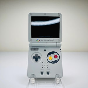 May include: A gray Nintendo Game Boy Advance SP handheld console, open and displayed on a clear stand. The screen is black. The console features a directional pad, buttons, and the text "SUPER Famicom".