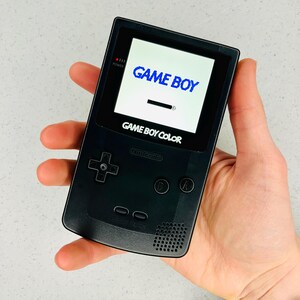 May include: A black Game Boy Color handheld video game console with a white screen displaying the text "GAME BOY" in blue. The console has a black and white button layout and a black and white Nintendo logo.