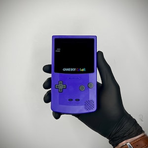 Può includere: Una console portatile per videogiochi di colore viola con schermo nero, caratterizzata dalla scritta GAME BOY COLOR in lettere multicolori, un logo Nintendo, un pad direzionale, pulsanti A e B, una spia di alimentazione e una griglia altoparlante.