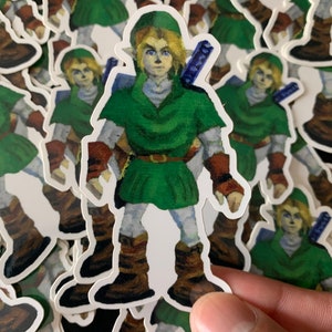 May include: A stack of stickers featuring a green-clad character with a sword. The character has blonde hair, green eyes, and a pointed hat. The stickers are printed on white paper.