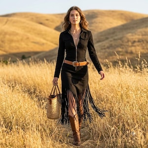 May include: A black long-sleeved dress with a front zipper closure, a tan leather belt with a silver buckle, and long fringe detailing along the hem. The dress is paired with brown leather cowboy boots and a woven straw tote bag with brown handles.