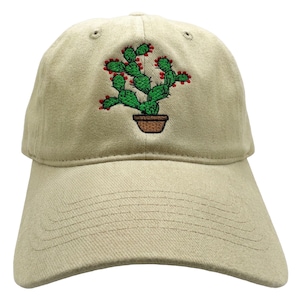 May include: A beige baseball cap featuring an embroidered design of a green prickly pear cactus in a brown pot. The cactus has small red accents on its pads. The cap has a curved brim and a structured crown with stitched eyelets on top.