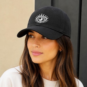 May include: A black baseball cap featuring a white embroidered graphic on the front. The design depicts an eye with a crescent moon pupil, surrounded by radiating lines and a row of small stars along the upper arch of the iris.