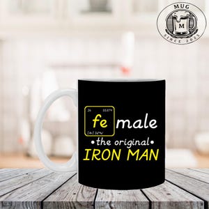 K&ouml;nnte beinhalten: Wei&szlig;er Keramikbecher mit schwarzem Hintergrund. Der Becher zeigt den Text "fe male &bull; the original &bull; IRON MAN" in Gelb und Wei&szlig; sowie das Eisensymbol aus dem Periodensystem.