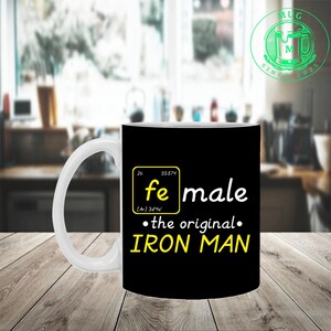 Puede incluir: Taza de cer&aacute;mica blanca con fondo negro que presenta la palabra "female" con el s&iacute;mbolo qu&iacute;mico del hierro y el texto "the original IRON MAN" en amarillo. La taza tiene un asa blanca y un logotipo verde.