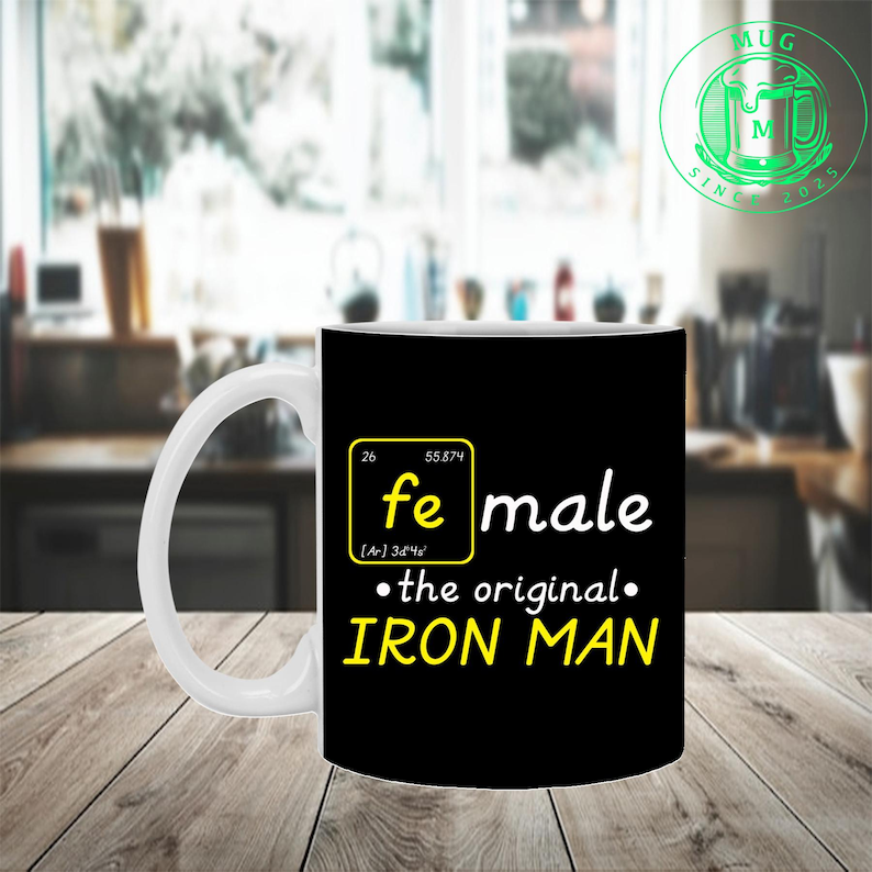 Puede incluir: Taza de cer&aacute;mica blanca con fondo negro que presenta la palabra "female" con el s&iacute;mbolo qu&iacute;mico del hierro y el texto "the original IRON MAN" en amarillo. La taza tiene un asa blanca y un logotipo verde.