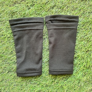 May include: Two black compression sleeves, possibly for athletic use, are shown against green grass. The sleeves are cylindrical, made of a perforated fabric, and have a folded top hem. The sleeves are designed to provide support and compression.