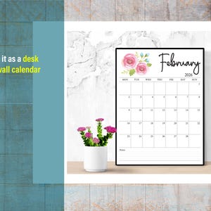 May include: A black framed calendar for February 2026, with floral accents. The calendar is displayed on a wooden surface, next to a small white pot with pink flowers. Text on the calendar includes the days of the week and dates.