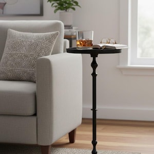 Puede incluir: Una mesa auxiliar negra y ornamentada con una tapa redonda, que sostiene un vaso de líquido ámbar con hielo, un libro abierto y gafas. La mesa tiene un tallo decorativo y una base circular. La mesa está junto a un sofá gris claro.