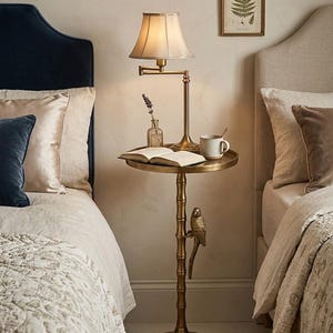 May include: A brass side table with a lamp, book, and mug. The table has a round top and a bamboo-style stem with a decorative bird. The lamp has a beige shade. The table is between two beds with pillows and bedding.