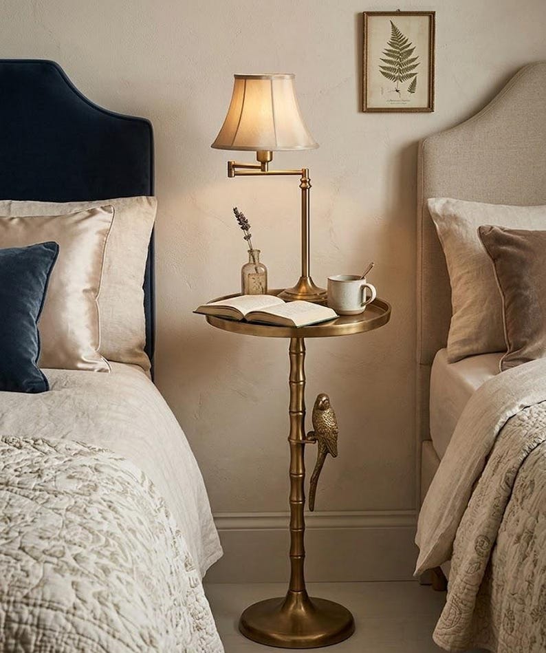 May include: A brass side table with a lamp, book, and mug. The table has a round top and a bamboo-style stem with a decorative bird. The lamp has a beige shade. The table is between two beds with pillows and bedding.