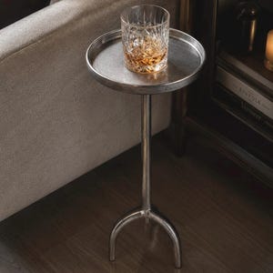 Puede incluir: Una mesa auxiliar redonda de metal plateado con una base de pedestal central que se divide en tres patas curvas. Un vaso transparente con un patrón de corte de diamante que contiene un líquido de color ámbar descansa sobre la mesa.