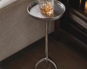 Può includere: Un tavolino rotondo in metallo color argento con una base a piedistallo centrale che si divide in tre gambe curve. Un bicchiere trasparente con motivo a taglio di diamante contenente un liquido color ambra poggia sul piano circolare.