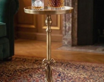 May include: A gold-colored side table with a round top, holding a crystal decanter and a glass with ice. The table has a decorative bow detail and a round base. The setting is a room with a fireplace and patterned rug.