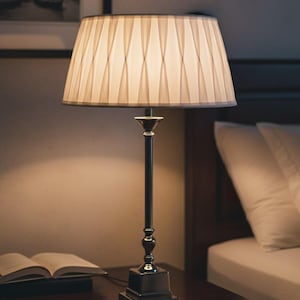 Op de afbeelding: Een bedlamp met een geplooide lampenkap. De lamp heeft een donkere metalen voet en steel en staat op een donker houten nachtkastje. Een open boek ligt naast de lamp. De achtergrond omvat een bed en een ingelijste foto.