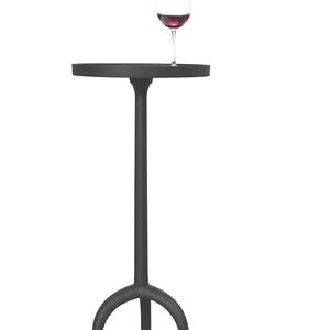 Może przedstawiać: Czarny, metalowy stolik boczny z okrągłym blatem i trójnożną podstawą. Na stole stoi kieliszek wina wypełniony czerwonym winem. Stół jest przeznaczony do użytku wewnątrz i na zewnątrz.