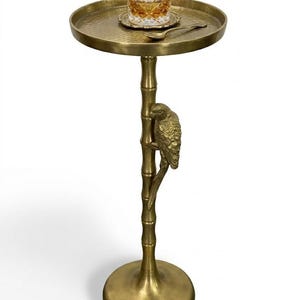 May include: A gold-colored side table with a round top and a parrot perched on a bamboo-style stem. A glass of amber liquid sits on the table, along with a small coaster and spoon. The table has a hammered texture.